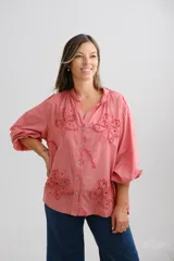 Blusa color coral de corte holgado, con cuello mao y cierre frontal de botones. Presenta mangas largas abullonadas y detalles de flores bordadas con técnica de calado en el frente.