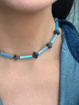 Collar corto de diseño artesanal compuesto por cuentas cilíndricas de vidrio en tono celeste y pequeñas piedras naturales esféricas en tonos azulados, intercaladas con detalles metálicos. Incluye cadena de acero quirúrgico para ajuste de largo.