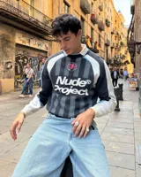 Suéter de punto con diseño inspirado en camisetas de fútbol retro. Presenta un patrón de rayas verticales en tonos gris oscuro, gris claro y celeste, con el logo de la marca en gran tamaño en el frente y un detalle de rosa bordada en el pecho. Posee cuello redondo y puños acanalados.