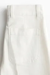 Jean de denim de algodón blanco con leve elasticidad, tiro alto y diseño de pierna ancha (wide leg). Cuenta con bolsillos de parche delanteros, botón metálico con diseño y pliegues marcados al frente y en la espalda.