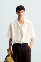 Camisa de calce relajado confeccionada en tejido de algodón color beige. Presenta un diseño de jacquard con líneas verticales desflecadas, cuello tipo bowling, manga corta y cierre frontal con botones.