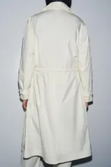 Gabardina oversize color blanco roto, con cuello solapa, manga larga y bolsillos laterales ocultos. Presenta cintura ajustable con cordones a tono y forro interior.