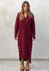 Vestido midi de punto color bordó, con cuello en V, mangas largas y diseño de rombos en relieve. Presenta aberturas laterales con botones decorativos y un corte asimétrico con el ruedo trasero más largo.
