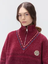 Pullover de punto con textura aterciopelada, diseño oversize, cuello polo con cierre frontal parcial y detalles de pespuntes en contraste. Presenta rayas horizontales en tonos tierra y un parche bordado con la letra M en el pecho.