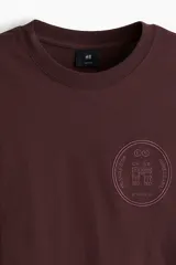 Remera de punto de algodón de peso medio color bordó, con cuello redondo ribeteado, hombros caídos y bajo recto. Presenta un diseño estampado circular en el pecho con texto y un arco. Corte holgado.