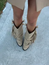 Botas de caña media confeccionadas en gamuza, con diseño holgado y detalle de flecos laterales con cadena metálica. Cuentan con suela plana y estilo western.