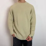 Sweater de tejido de punto en color beige, con cuello redondo y mangas largas con puños acanalados. Presenta un diseño holgado y una etiqueta pequeña con el logo de la marca en el puño izquierdo.