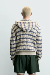 Cárdigan de punto con diseño de trenzas y rayas horizontales en tonos beige y azul. Presenta corte relajado, capucha, cierre frontal con cremallera y puños y ruedo en tejido rib.