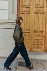 Cazadora estilo bomber color marrón oscuro, con efecto piel, cuello subido y manga larga. Tiene bolsillos delanteros con cierre de cremallera y acabados elásticos en puños y bajo. El cierre frontal es con cremallera y botones a presión ocultos.