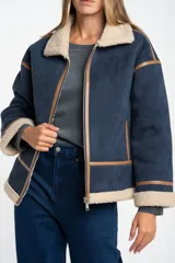 Campera tipo gamulán de corte oversize, color azul marino con interior y cuello de corderito en tono beige. Presenta detalles de costuras y bordes en cuero sintético color marrón, cierre frontal y mangas largas.