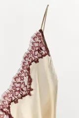 Vestido mini tipo lencero de tejido satinado color crema, con escote en pico y tirantes finos. Presenta detalles de encaje a contraste en color bordó en el escote y en el ruedo.