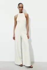 Mono largo de color blanco con cuello halter y espalda descubierta. Presenta un diseño de pierna ancha y cierre posterior con cremallera oculta y botones.