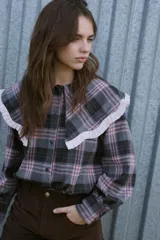 Camisa de manga larga con estampado de cuadros en tonos grises y rosados. Presenta un cuello grande estilo babero con volados en color rosa claro y cierre frontal con botones.