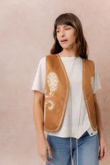 Chaleco corto de efecto gamuza en color tostado, con bordados decorativos en contraste y terminaciones bordadas en los bordes. Presenta escote en V y diseño recto.