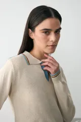 Remera de punto color crudo con cuello redondo y detalles de rayas en azul, amarillo y bordó. Presenta un parche bordado en el pecho con el logo de Manos del Uruguay y la inscripción 2026.