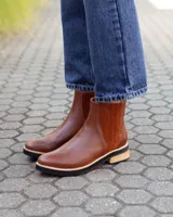 Botinetas de cuero color marrón con diseño de paneles combinados, incluyendo una sección lateral con textura grabada. Presenta una suela plana con detalle de madera y base de goma negra.