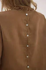 Blusa de lino color marrón, de corte recto y mangas 3/4. Presenta escote redondo con pequeños volados y pliegues frontales, además de detalles de encaje en las mangas y puños con volados.