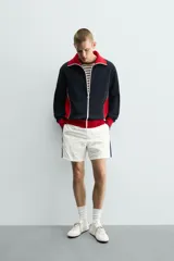 Chaqueta deportiva de corte regular, confeccionada en tejido piqué con interior forrado en red. Presenta un diseño color block en negro y rojo con detalles en blanco, cuello subido, cierre frontal con cremallera y bolsillos laterales con cierre.