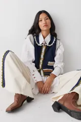 Chaleco de lana color marrón con cuello estilo bebé, cierre frontal con botones grandes y detalle de puntilla en cuello, sisas y bolsillo frontal. Presenta una franja decorativa en color rojo en el ruedo.