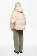 Chaqueta acolchada tipo puffer de corte globo en color beige, con cuello subido, capucha, cierre frontal con cremallera y botones de presión, y bolsillos delanteros con tapeta.