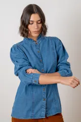 Camisa de denim azul con cuello alto tipo mao con volados, mangas largas abullonadas y cierre frontal con botones metálicos.