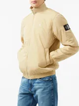 Campera de nailon color beige con cierre frontal de cremallera, cuello alto de canalé, puños y bajo elásticos. Presenta el logo de Calvin Klein bordado en el pecho y un parche con el logo en la manga izquierda.