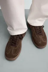 Zapatilla de estilo urbano confeccionada en piel con acabado de serraje en color marrón. Presenta cierre mediante cordones al tono, detalle de pespuntes en el empeine y suela de goma a tono.