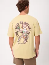 Remera de manga corta color amarillo, corte holgado, con gran estampado en la espalda de un perro en bicicleta con una tabla de surf y un loro, acompañado del texto "Wild by Nature Rusty Worldwide".