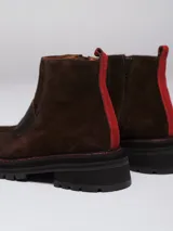 Botinetas de cuero y gamuza en tono marrón oscuro, con diseño de puntera reforzada y costuras decorativas en color rojo. Presentan una banda ancha sobre el empeine, cierre lateral y una lengüeta roja en el talón para facilitar el calce.