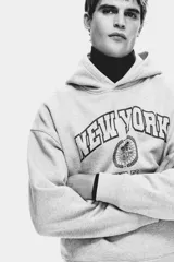 Canguro H&M de mezcla de algodón en color gris melange, con capucha de doble capa, bolsillo tipo canguro y hombros caídos. Presenta un estampado frontal con la inscripción "NEW YORK PHYS. ED." en tonos azules y rojos con detalles de escudo.