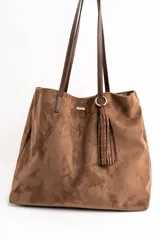 Bolso tipo tote de gran tamaño confeccionado en gamuza sintética color tostado. Presenta un diseño minimalista con asas largas de hombro y una pequeña placa metálica frontal con el logo de la marca.