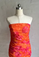 Vestido corto de microtul con diseño drapeado, escote strapless y estampado floral en tonos naranja y fucsia.