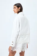 Sobrecamisa blanca de corte recto con cuello clásico, cierre frontal de botones y bolsillos delanteros. Presenta un delicado detalle de bordado inglés en el ruedo.