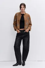 Chaqueta de gamuza color tostado con diseño de flores bordadas con pedrería metálica y cristales. Presenta cuello redondo, cierre frontal con botones a presión y mangas largas.