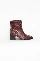 Bota corta con punta afilada y taco cuadrado, confeccionada en material con textura de piel de serpiente en tonos bordó y negro. Presenta una correa decorativa con hebilla metálica dorada en el lateral.
