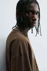 Camiseta de algodón color marrón oscuro, corte relajado (relaxed fit) y manga corta. Presenta un estampado frontal con un lobo o cánido corriendo, el texto "The Riders Project", "VISION & TEMPER" y "0.79 per gallon" en tonos grises y rosados.