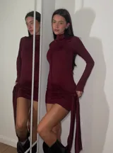 Vestido corto de cuello alto y manga larga, confeccionado en tejido elástico color bordó. Presenta un diseño ajustado al cuerpo con un detalle de nudo lateral y caída de tela asimétrica.