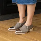 Zapato tipo mocasín de cuero vacuno en tono gris topo, con puntera reforzada en cuero metalizado color bronce. Presenta un diseño de corte lateral y suela plana con pequeña elevación en el talón.