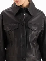 Chaqueta de cuero ovino color negro, con textura graneada, silueta de hombros caídos y largo a la cadera. Presenta recortes en el delantero y la espalda, dos bolsillos superiores con tapa y dos bolsillos laterales, cierre frontal con botones metálicos y cuello camisero.