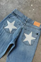 Jean de mezclilla celeste, corte holgado o recto, con grandes estrellas estampadas en color blanco o gris claro en los bolsillos traseros. Tiene un parche de cuero marrón en la cintura trasera con el logo de la marca.