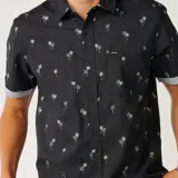 Camisa Rip Curl de manga corta, color negro con estampado de pequeñas palmeras blancas, cuello camisero, cierre frontal de botones y un bolsillo ojal en el pecho.