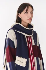 Bufanda de punto con capucha integrada, diseño a rayas en tonos azul marino, bordó y crema. Presenta un borde con detalle de costura en color crema y una etiqueta rectangular bordada con el nombre de la marca en uno de sus extremos.