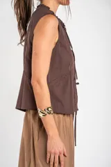 Blusa de algodón sin mangas en color blanco off-white, con cuello alto con volados y cierre frontal de cordones ajustables con lazos. Presenta detalles de encaje y bordados sutiles en la parte delantera.