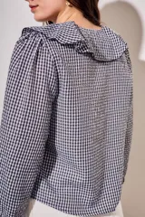 Blusa de corte oversize con estampado de cuadros vichy en blanco y negro. Presenta un cuello con volados, mangas largas con puños elásticos y un cierre frontal con lazo negro.