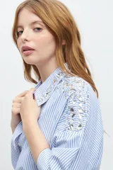 Camisa celeste a rayas verticales blancas, con cuello clásico, manga larga y bordados de piedras brillantes en hombros y cuello.