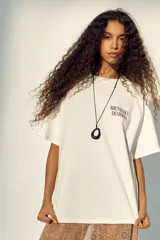 Remera oversized de punto suave de algodón, color blanco, con cuello redondo acanalado, hombros caídos y diseño estampado en el frente con la frase 'Arenas del Desierto'.