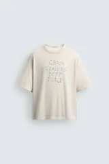 Camiseta de corte holgado y estilo cropped, color beige con efecto lavado. Presenta cuello redondo, mangas cortas y una estampa frontal con texto en azul que simula trazos de rotulador.