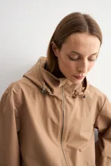 Trench color camel con capucha desmontable, cierre frontal con cremallera, bolsillos laterales y mangas largas con ajuste en puños.