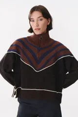 Buzo de tejido de punto con diseño oversized y corte a la altura de la cadera. Presenta cuello alto con cierre frontal, mangas largas y un patrón geométrico en V en la parte superior que combina tonos marrones, azul y detalles en blanco.