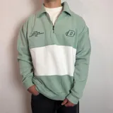 Buzo de algodón con medio cierre frontal y cuello tipo polo. Presenta un diseño de bloques de color en tonos gris oscuro y blanco, con bordados decorativos en el pecho.